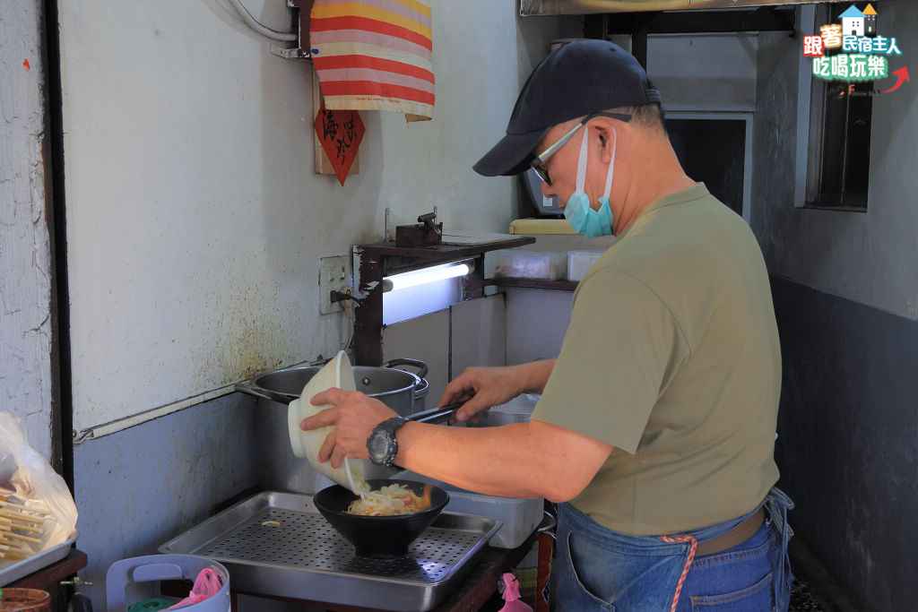 老闆在窄小廚房裡俐落地將配料夾進黑色碗中備料的畫面